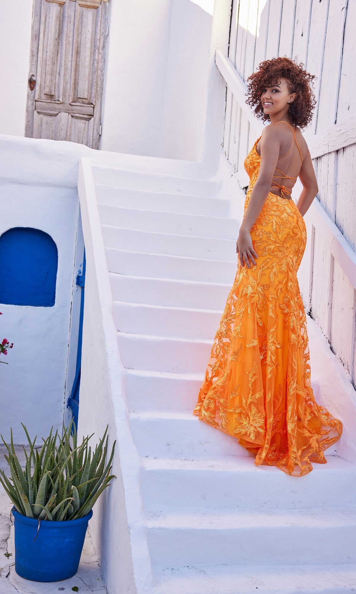 A woman in the Ellie Wilde Long Designer Formal Dress EW35109 stands on white outdoor stairs, glancing over her shoulder; a potted plant and blue accents complete the setting.